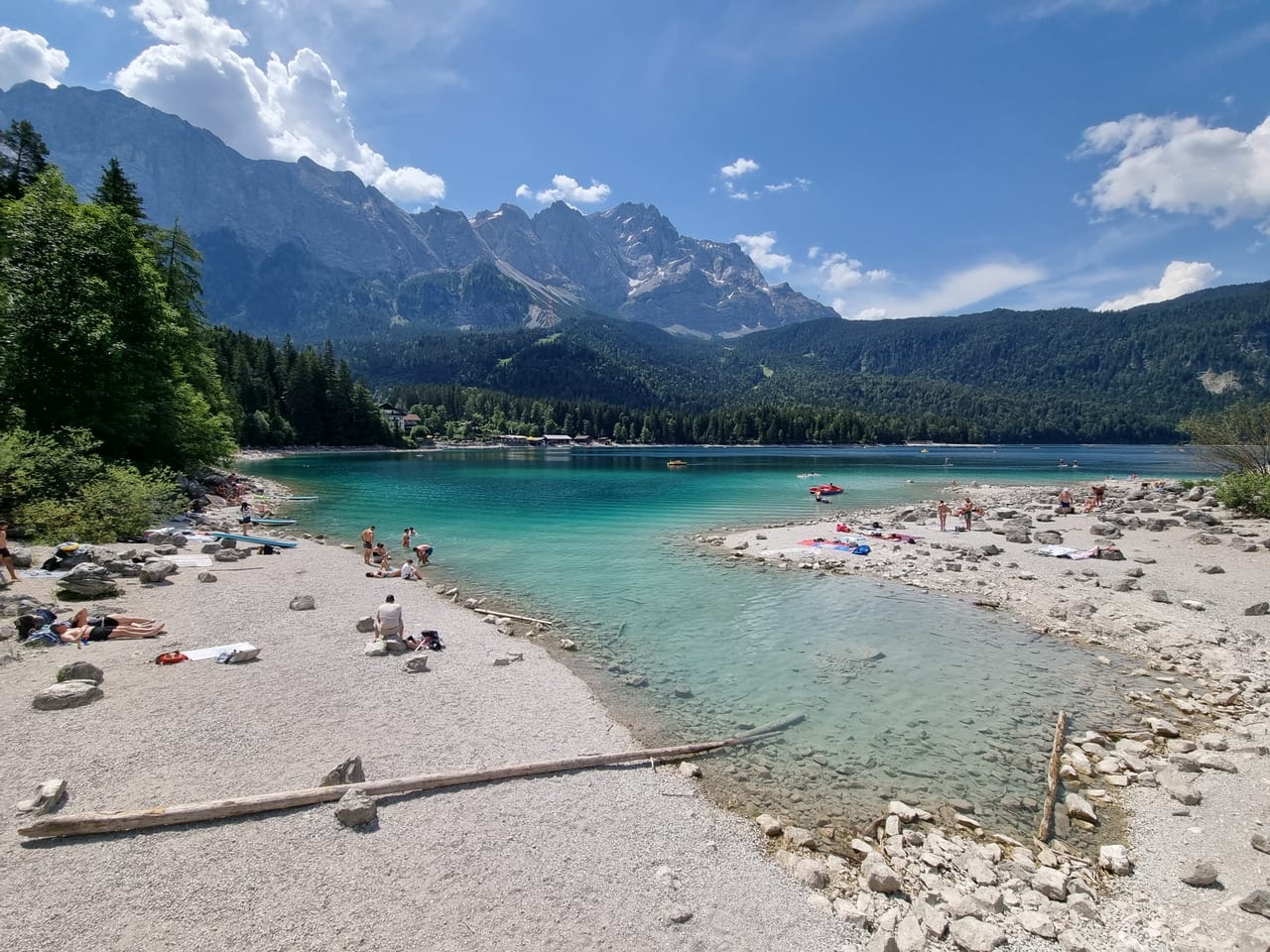 The turquoise water of Lake Eibsee with a pebbled shore in the foreground and the Zugspitze massif under a blue summer sky in the Bavarian Alps